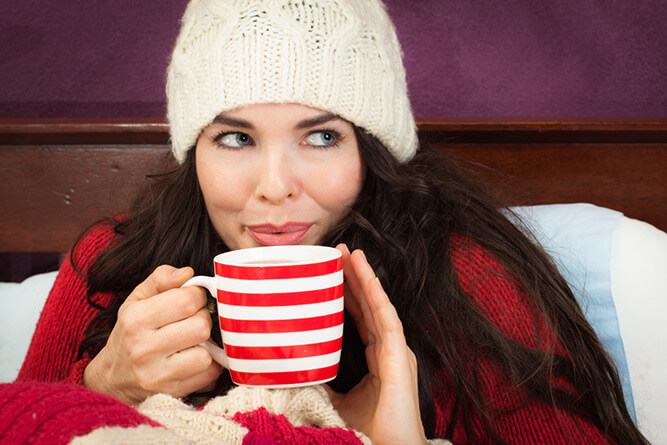 photo of a woman in bed drinking tea wearing a warm hat and jumper