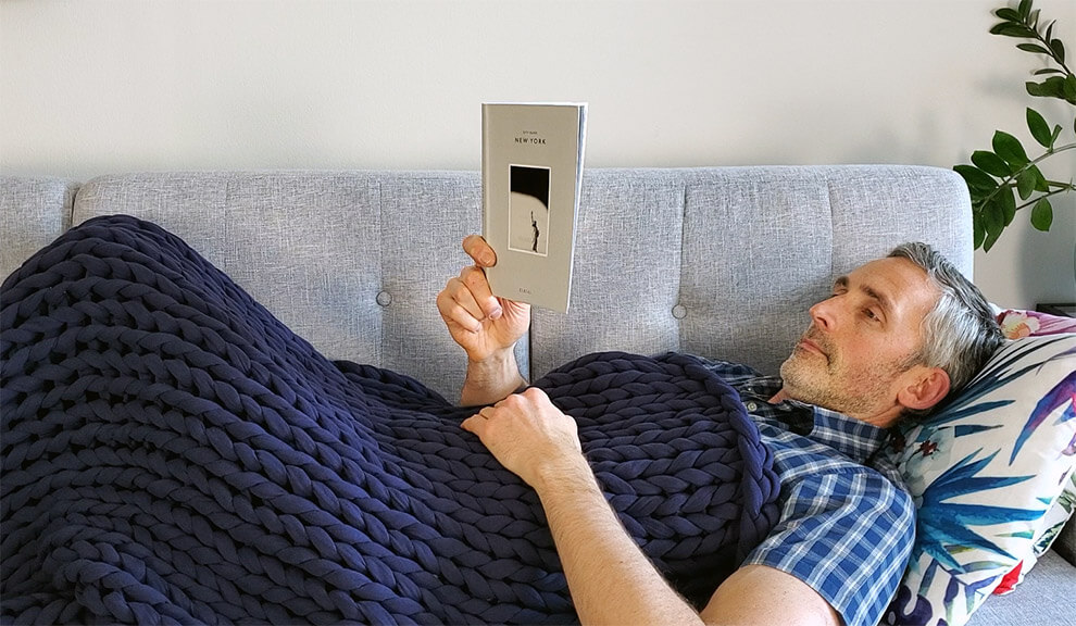 photo of a man reading a book and relaxing under a weighted blanket on the sofa